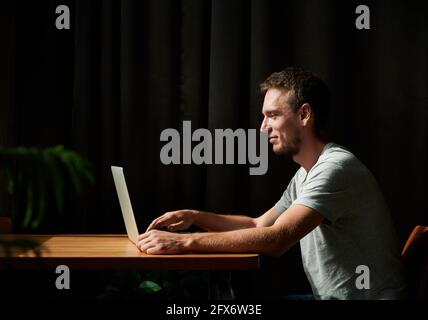 Side view of male student sitting by table and typing on laptop. Young man in grey T-shirt is working on laptop at home and smiling. Concept of studying from home. Stock Photo