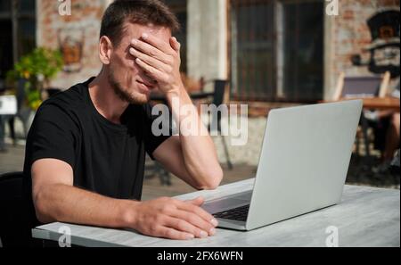 Dissapointed male with hand on face tormented by work. Young man is working on laptop in cafe. Concept of hard worker. Stock Photo