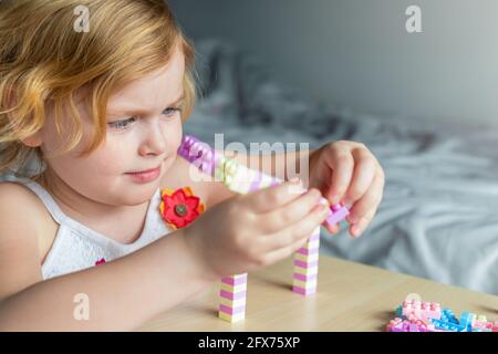 Little beautiful girl playing with toy plastic building blocks, sitting at the table. Small child busy with fun creative leisure activity. Development Stock Photo