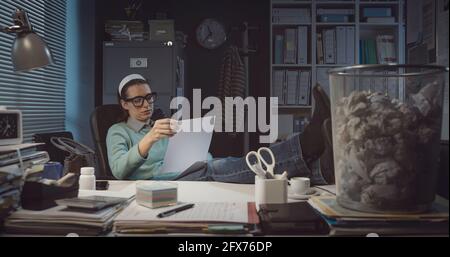 Frustrated woman with feet on desk reading paperwork late at night, trash can filled with crumpled paper in the foreground Stock Photo