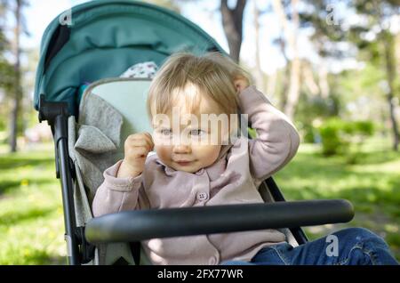 Baby in stroller on a walk in spring park. Adorable little girl in common clothes sitting in blue pushchair. Child in buggy Stock Photo