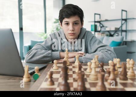 Boy learning chess at home, he is using a laptop and staring at the chessboard Stock Photo