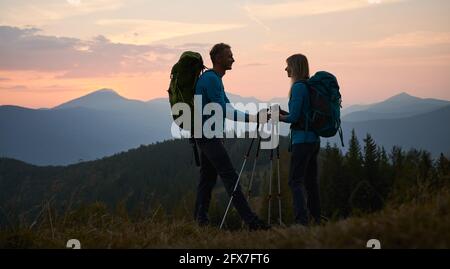 Full length of smiling man and woman travelers hiking together in mountains. Silhouette of happy young couple with backpacks standing on grassy hill with sunset and grassy hills on background. Stock Photo