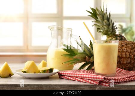Glass and jug with pineapple juice with milk and pineapple portions on wooden table and window background. Stock Photo