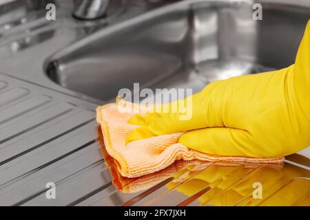 Hand in yellow latex protective glove wiping stainless steel sink with microfiber cloth. Household cleaning tools. Stock Photo