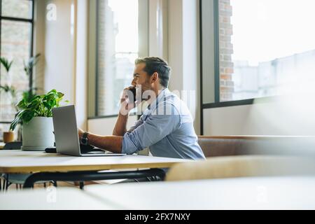 Man in office talking on mobile phone. Young male at office desk with laptop speaking on smart phone. Stock Photo