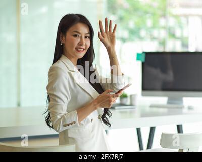 Portrait of businesswoman greeting, raised hand to say hi and smiling to camera while using smartphone in office room Stock Photo