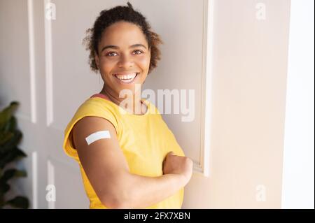 Cheerful vaccinated African-American woman showing arm with medical patch and laughs, black female getting vaccine dose against covid, plaster on her shoulder, isolated on white. Healthcare concept Stock Photo