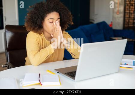 Overworked African-American freelancer woman feels eyes strain due to long-term work with a laptop, tired dark skinned female sits at the table with closed eyes, feeling stressed. Overload concept Stock Photo
