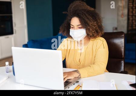 African young woman sits at the desk wearing protective mask against coronavirus and flu using laptop computer in the home office, working remotely with face covered Stock Photo