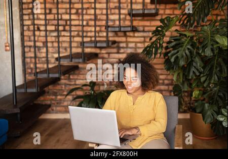 Attractive happy young African-American female entrepreneur using laptop for remote work, sitting in a cozy loft apartment, black woman enjoys freelance work, designer works on the distance Stock Photo