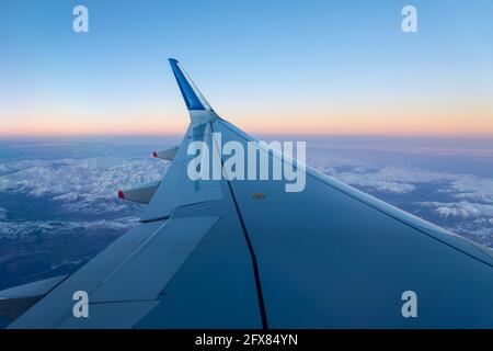 Airplane wing in flight over the Alps in winter at sunset, travel concept Stock Photo