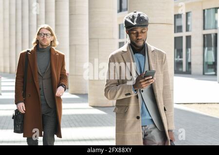 African young man using mobile phone while walking along the street with man in the background Stock Photo