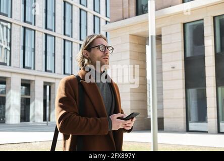 Young man in eyeglasses walking along the street and using mobile phone Stock Photo