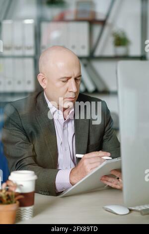 Concentrated mature analyst with bald head sitting at desk and working with data in office Stock Photo