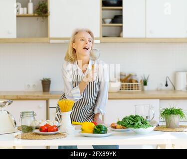 Playful carefree senior woman in apron using metal strainer as microphone and singing, having fun while cooking healthy food in modern light kitchen at home. Happy mature female enjoying retired life Stock Photo