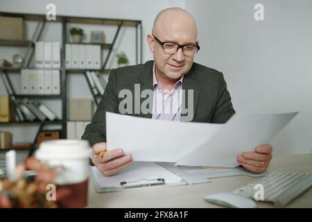 Content bald mature businessman in glasses sitting at office table and reading papers in office Stock Photo
