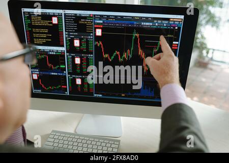 Over shoulder view of stock market analyst pointing at computer monitor while working with colorful graph Stock Photo