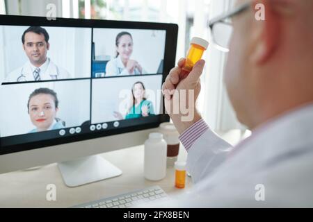 Over shoulder view of doctor sitting at table and showing pill bottle to web camera while discussing new medicine with colleagues online Stock Photo