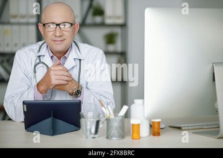 Portrait of content confident mature Caucasian doctor in white coat sitting at table with pills and computer and using tablet Stock Photo