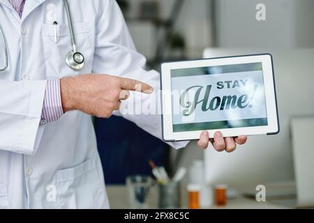 Close-up of unrecognizable doctor in white coat pointing at tablet with Stay home text Stock Photo