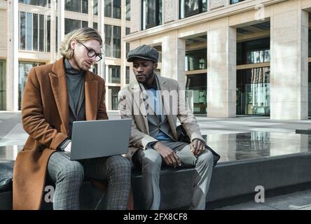 Two young colleagues using laptop and discussing online work together outdoors Stock Photo