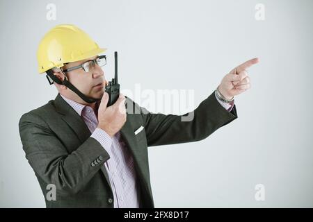 Serious mature building contractor or engineer in work helmet and glasses using walkie-talkie while explaining project plan to worker Stock Photo