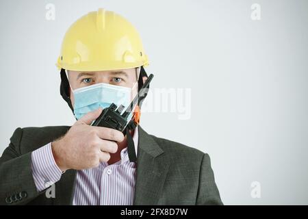 Portrait of mature building contractor in yellow hardhat and mask using walkie-talkie while transmitting message Stock Photo