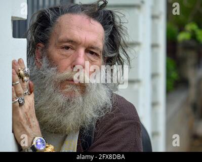 Old emotional dignified Caucasian man with thick beard and disheveled hair hides behind a wall, looks at the viewer and shows the rings on his hand. Stock Photo