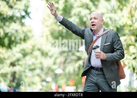 Concerned bald mature businessman in suit wearing bag over shoulder waving hand and catching taxi while being late for work Stock Photo