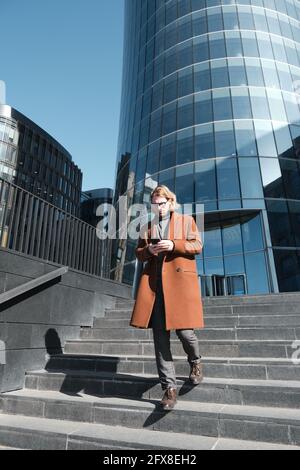 Young businessman using mobile phone while moving down the stairs with modern building in the background Stock Photo