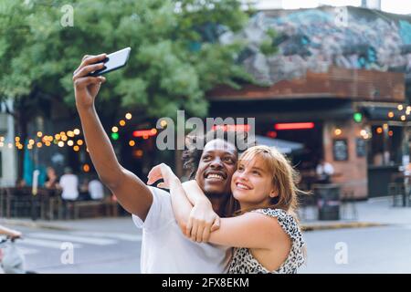 Happy young multi-ethnic couple taking selfies on the street in the evening. Stock Photo