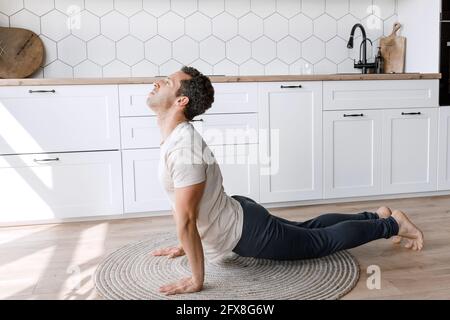 Focused guy doing yoga exercises during home training. Concentrated man doing cobra pose at carpet on the floor in modern kitchen, relieve stress Stock Photo