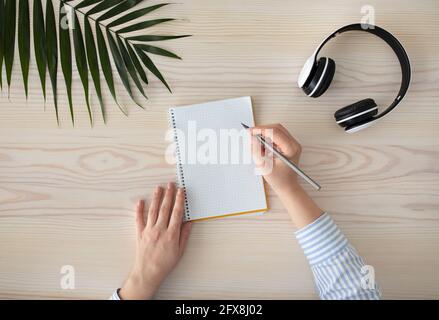 To do list. Female hands writing with pencil in notebook , sitting on wooden table with headphones and tropical leaf Stock Photo