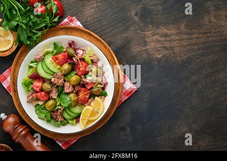 Tuna salad with fresh vegetables, olives, capers and lemon served in bowl on old wooden background. Top view with copy space Stock Photo