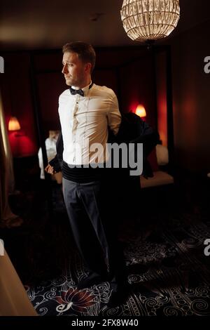 Morning of the groom before the wedding. Groom morning preparation. Young and handsome groom getting dressed in a wedding bow tie. Stylish man in suit Stock Photo