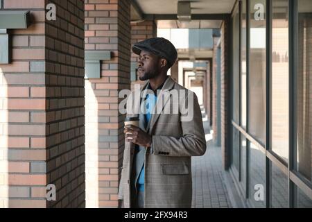 African young businessman holding cup of coffee and looking away standing on the street Stock Photo