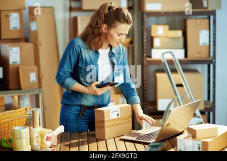 Delivery business. happy modern female in jeans with laptop and parcels using smartphone applications in the warehouse. Stock Photo