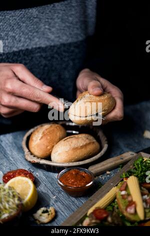 a young caucasian man cuts a bread bun to prepare a sandwich or some vegan snacks, sitting at a gray wooden table, next to a tray with some vegan appe Stock Photo