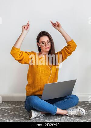 Young woman listening music headphones and dancing, using laptop sitting on carpet on white background Stock Photo