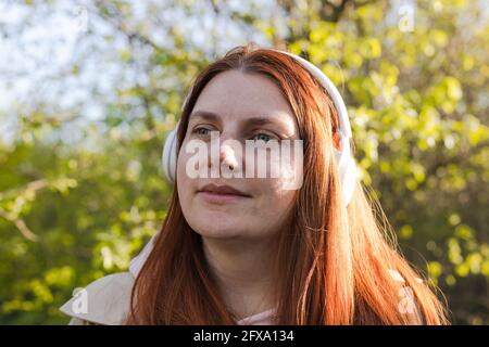 Beautiful caucasian young woman listening music with headphones on sunny day while walking in city park Stock Photo