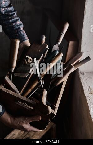 From above anonymous artisan carrying wooden crate with assorted instrument near window in dark carpenter workshop Stock Photo