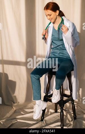 Portrait of smiling charming female doctor in white coat sitting on chair near window in sunny day in medical clinic office. Stock Photo