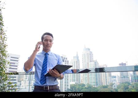 Smart Asian Business man using smart phone and document to working on window with city building and green balcony Stock Photo