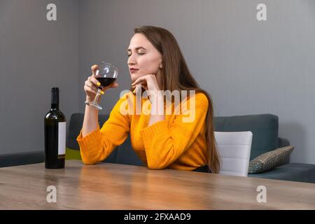 Beautiful young dark-haired woman sits at a large table in the living room and inhales the aroma of red wine from a glass Stock Photo