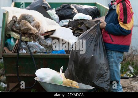 Young man in s jacket throws out a large plastic garbage bag to the garbage countryside container outdoors Stock Photo