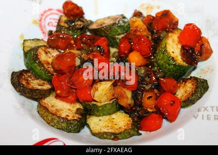 Close-up of fried zucchini and cherry tomatoes, with shredded parmesan cheese, basil, and seasoning, in a white serving dish. Stock Photo