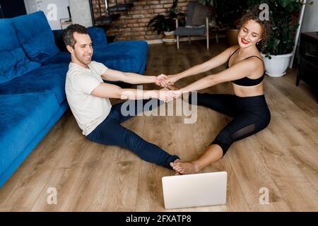 Healthy couple stretching watching fitness video tutorial online on laptop. Young smiling fit man and woman doing yoga workout at home sitting on Stock Photo
