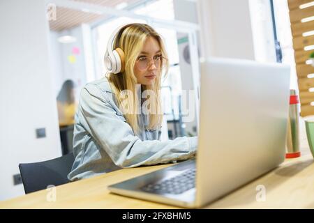 Blond businesswoman using laptop with headphones in office Stock Photo