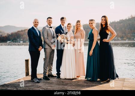 Bride and groom standing along with friends on jetty over lake Stock Photo